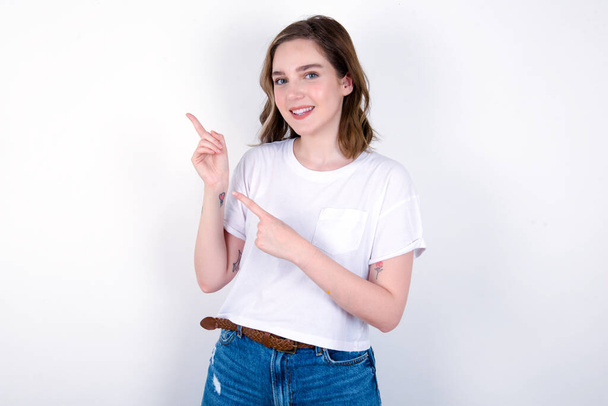 Positive girl with beaming smile pointing with two fingers and looking on empty copy space. Advertisement concept. Young caucasian woman wearing white T-shirt over white background  - Photo, Image