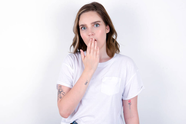 Oh! I think I said it! Close up portrait girl covering open mouth by hand palm, looking at camera with big eyes.  Young caucasian woman wearing white T-shirt over white background  - Photo, Image