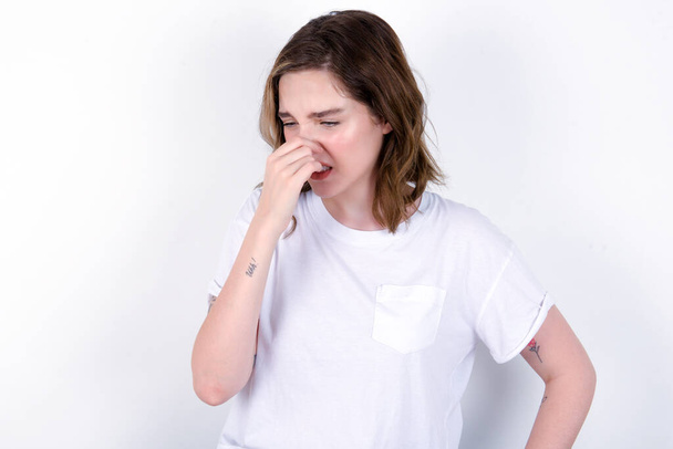 chica sosteniendo su nariz debido a un mal olor. Mujer joven caucásica vistiendo camiseta blanca sobre fondo blanco  - Foto, Imagen