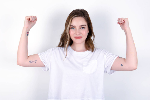 Waist up shot of girl raises arms to show muscles feels confident in victory, looks strong and independent, smiles positively at camera. Sport concept. Young caucasian woman wearing white T-shirt over white background  - Photo, image