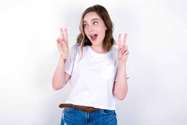 Isolated shot of cheerful girl makes peace or victory sign with both hands, feels cool. Young caucasian woman wearing white T-shirt over white background  - Photo, Image