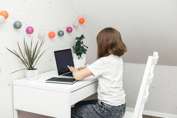 A teenage girl is sitting at a table in her room and working on a laptop, watching webinars or chatting with friends. The role of gadgets in the life of modern children - Foto, Imagem