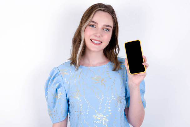 Χαμογελαστή κοπέλα που δείχνει άδεια οθόνη τηλεφώνου. Έννοια διαφήμισης και επικοινωνίας. Νεαρή λευκή γυναίκα με μπλε μπλουζάκι πάνω από λευκό φόντο  - Φωτογραφία, εικόνα