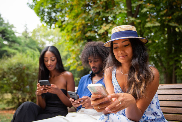 Three friends sitting on a bench in a park use their smartphones, multi-ethnic group. - Photo, image
