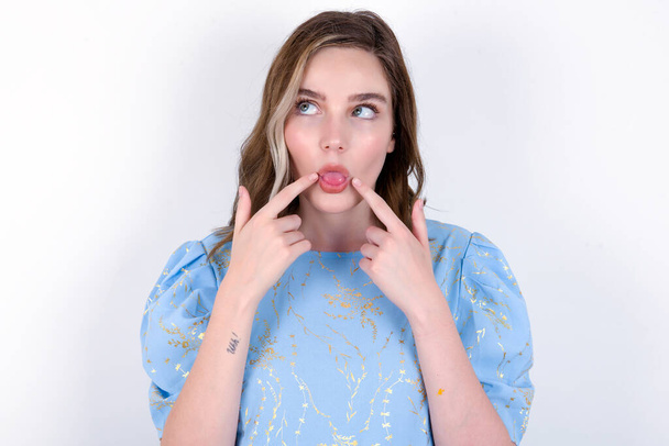молода біла жінка в блакитній футболці на білому тлі, що показує язик, смішна похмура - Фото, зображення