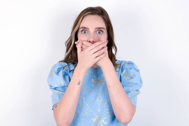 Verbaasde jonge blanke vrouw draagt blauw T-shirt over witte achtergrond dekt mond met beide handen bang of horen verbluffende roddels. - Foto, afbeelding