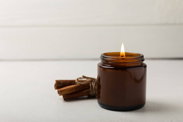 A cozy burning candle in a brown glass jar with cinnamon sticks on a white wood texture. winter home decor. Soy ecological candle. - Fotoğraf, Görsel