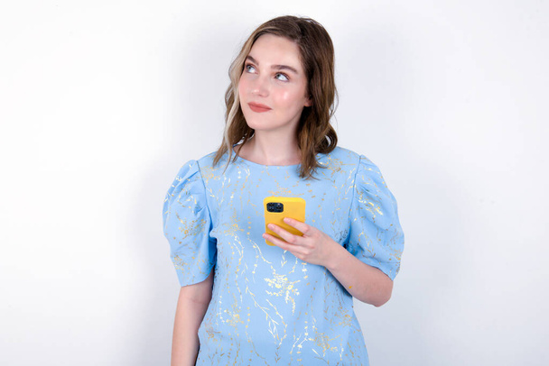 白い背景の上に青いTシャツを着ている若い白人女性は携帯電話を持ち,ソーシャルネットワークのための高速インターネットを使用します. オンラインコミュニケーションと現代技術のコンセプト - 写真・画像