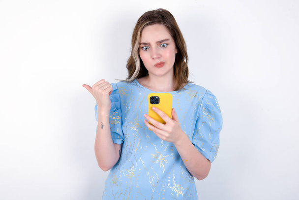 młoda kaukaska kobieta w niebieskiej koszulce na białym tle punkty kciuk dalej i pokazuje puste miejsce na bok, trzyma telefon komórkowy do wysyłania wiadomości tekstowych. - Zdjęcie, obraz