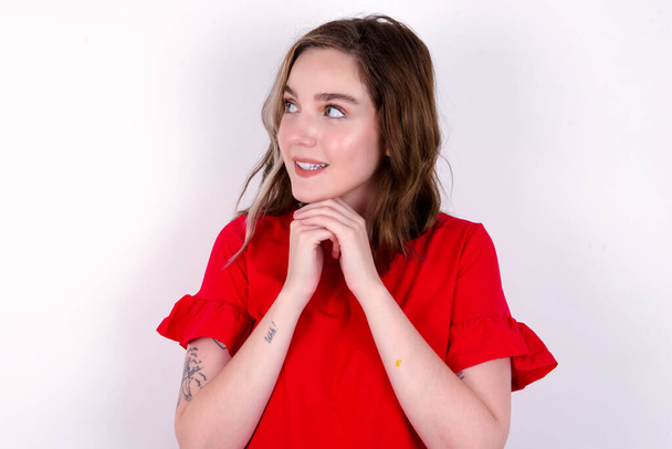 Happy young caucasian woman in red t-shirt over white background anticipates something awesome to happen, looks happily aside, keeps hands together near face, has glad expression. - Photo, Image