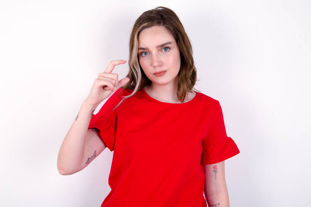 Неймовірна молода біла біла жінка в червоній футболці на білому тлі демонструє знак невеликого розміру, демонструючи щось не дуже велике. Концепція мови тіла. - Фото, зображення