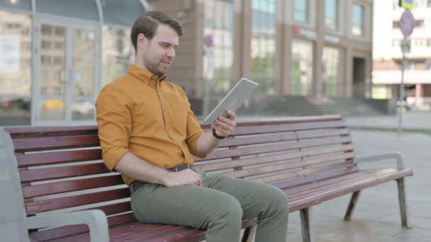 Online Video Chat on Tablet by Casual Young Man Sitting Outdoor on Bench  - Filmmaterial, Video