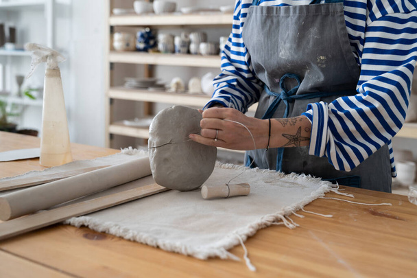Ceramic craft master building pieces out of clay by hand, close up of female potter wearing apron using wire clay cutter trimming tool, cropped photo, making handmade pottery in studio. Small business - Fotografie, Obrázek