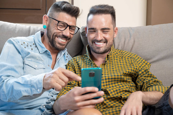Щаслива молода пара гей, використовуючи мобільний телефон, сидячи на дивані у вітальні. Високоякісна фотографія
. - Фото, зображення