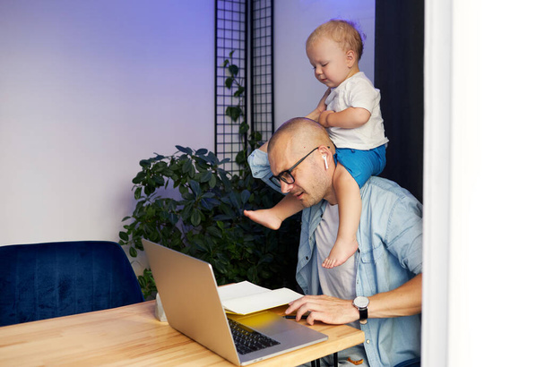 Young father working remotely on laptop with little baby son in his arms - Foto, Bild