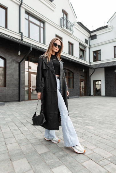Stylish beautiful young girl with sunglasses in fashionable outfit with a leather-length coat, flare high-waisted jeans and sneakers with a purse walking on the street - Foto, Imagen