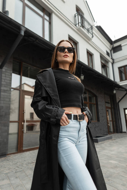 Beautiful fashionable woman model with cool black sunglasses in stylish street clothes with a trendy leather coat, tank top and high waisted jeans in the city - Foto, afbeelding