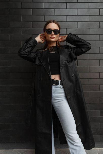 Stylish glamorous young pretty woman model in fashionable black clothes with a fashion long leather coat with a black T-shirt and vintage jeans standing near a black brick wall - Foto, imagen