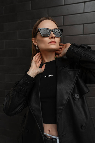 Fashion urban female portrait of beautiful girl with trendy sunglasses in black leather coat with black t-shirt stands and poses near a black brick background. Female style outfit , beauty and fashion - 写真・画像