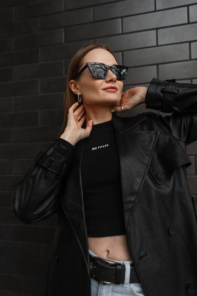 Modieus mooi stijlvol hippe meisje in stijlvolle zonnebril in trendy urban casual kleding met een zwarte leren jas en een zwart mockup t-shirt poseert vlakbij een zwarte bakstenen muur - Foto, afbeelding