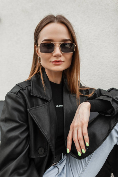 Women's street fashionable portrait of a young beauty girl with trendy gold eyeglasses in fashion black outfit with a stylish leather coat and T-shirt sitting on the street - Fotó, kép