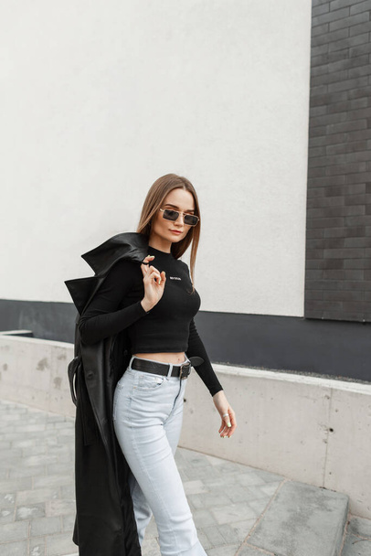 Beautiful stylish urban model woman with gold vintage eyewear in fashionable urban casual clothes with black T-shirt and high waisted jeans with coat walking on the street near the building - Φωτογραφία, εικόνα