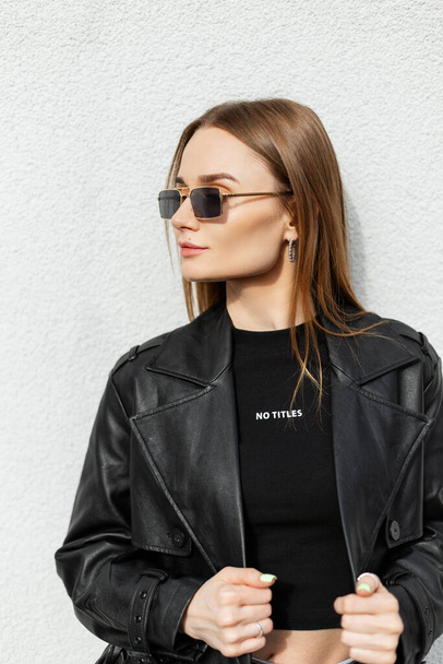 Beautiful young pretty woman model with fashionable sunglasses in black clothes with stylish leather coat and fashion black t-shirt stands near a white wall on sunny day - Foto, imagen