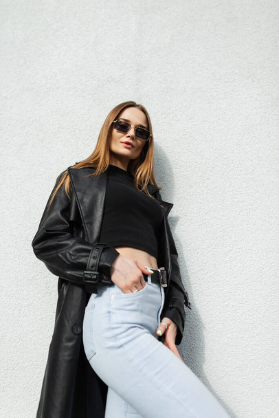 Fashionable beautiful young woman model with gold vintage sunglasses in a fashion long black leather coat with T-shirt and high-waisted jeans standing near the white wall on the street - Foto, imagen