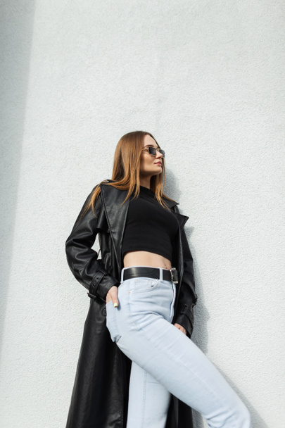 Glamour fashion street woman with vintage sunglasses in stylish black leather long coat with t-shirt and blue jeans stands near a white wall on the street - Fotoğraf, Görsel