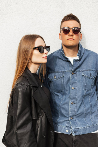 Cool street portrait of a beautiful young couple with vintage sunglasses wearing trendy casual clothes with a denim jacket and leather coat stand near the wall. Fashionable girl and stylish man - 写真・画像