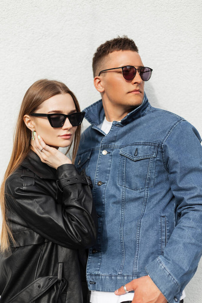 Stylish beautiful youth couple woman and a guy with cool sunglasses in fashionable casual clothes with a jacket, T-shirt and jeans standing on a white background on the street - Foto, Bild