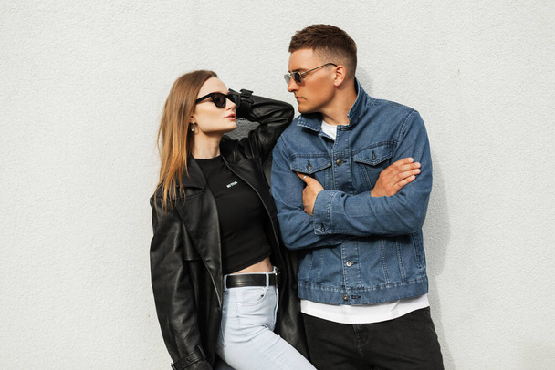 Fashionable couple beautiful model woman and handsome stylish man with vintage sunglasses in casual urban clothes with leather black coat and denim jacket poses near the wall on the street - Foto, immagini