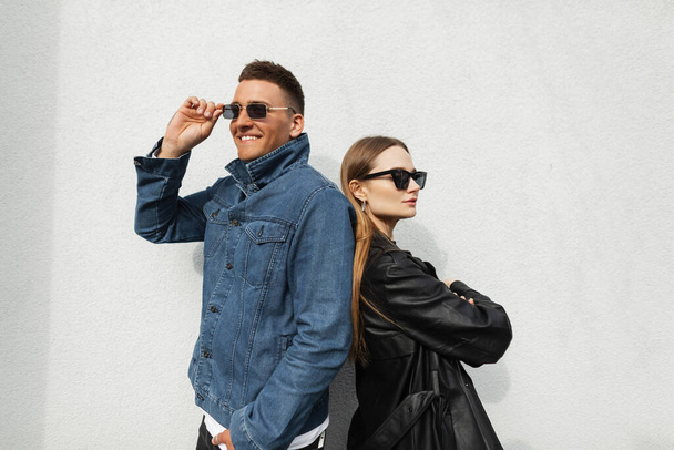Fashionable young couple in trendy urban casual clothes posing near a wall on the street. Fashion handsome happy hipster man and his girlfriend wearing vintage sunglasses in stylish denim outfit - Photo, image