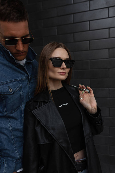 Fashionable young beauty girl with fashion eyewear in black trendy outfit with bag stands near a black brick wall with cool hipster man - Photo, image