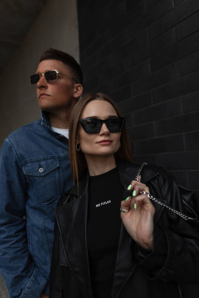 Fashionable beautiful couple in stylish street fashion outfit with jacket, bag and sunglasses walking on the street on a dark background. Woman's and man's urban casual outwear - Photo, image