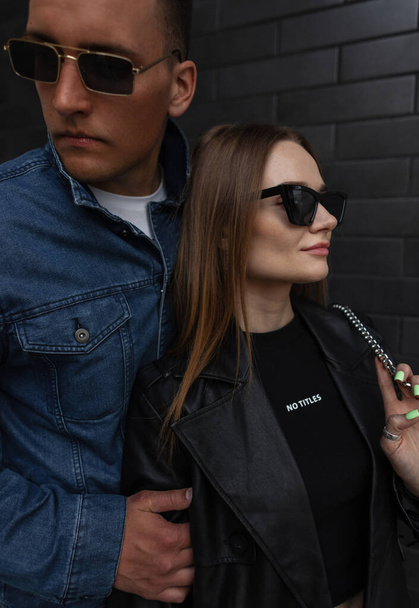 Modieuze urban mooi koppel man en mooie vrouw in stijlvolle denim outfit met jas en trendy zonnebril poseert in de buurt van een zwarte bakstenen muur op de straat - Foto, afbeelding