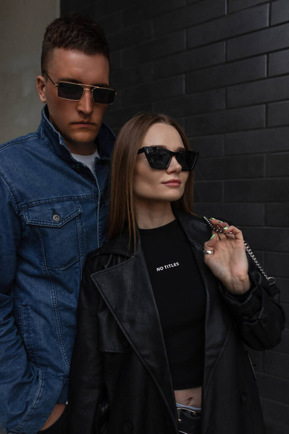 Divatos utcai fiatal pár gyönyörű lány és jóképű férfi divatos ruhák napszemüveg és táska sétálnak közel a fekete tégla fal a városban - Fotó, kép