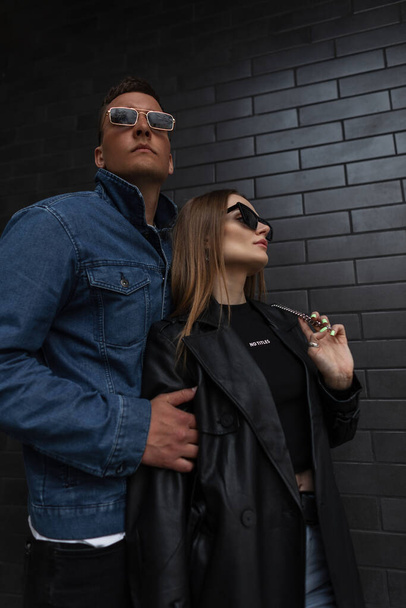 Cool beautiful fashionable glamorous hipster couple in stylish clothes with sunglasses stands near a black brick wall on the street. Beautiful model girl and handsome guy in trendy casual outwear - 写真・画像