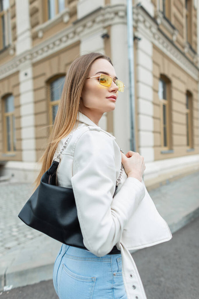 Fashion straat mooi meisje in stijlvolle kleding met lederen jas, portemonnee en gele bril wandelen in de stad - Foto, afbeelding