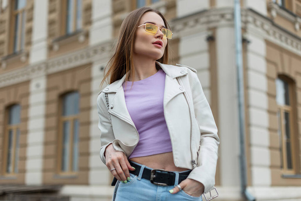 Fashionable beautiful young woman model with yellow sunglasses in a fashionable leather jacket and purple T-shirt walking in the city - Fotografie, Obrázek