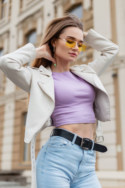 Stylish beautiful young hipster girl with yellow fashion glasses in a trendy white leather jacket with a purple T-shirt and fashion high waist jeans poses in the city - Foto, Imagen