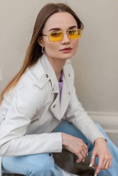Stylish beautiful young hipster woman with yellow sunglasses in a fashionable leather jacket and jeans sits near a wall on the street - Photo, Image