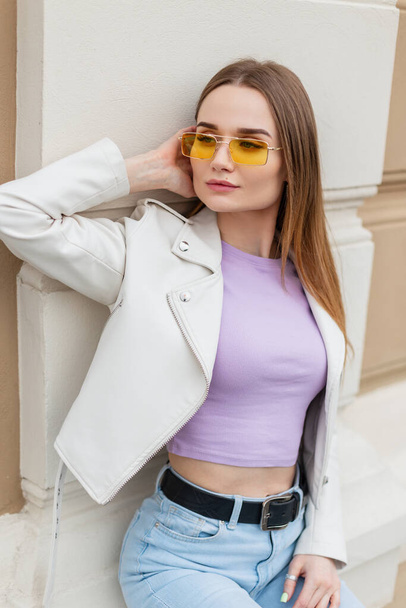 Trendy hipster beautiful young woman model in fashionable bright casual street clothes with a leather jacket, purple T-shirt and jeans sits near a vintage building on the street - Fotó, kép