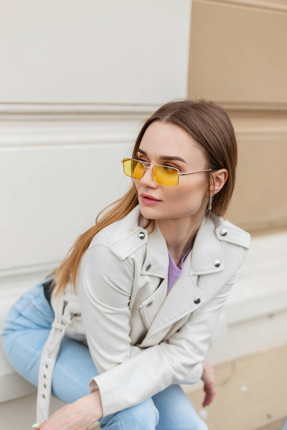 Fashion hipster woman model with yellow sunglasses in white leather jacket and blue jeans sits near a beige wall on the street - Foto, Bild