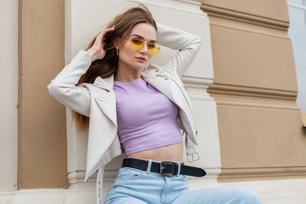 Stylish beautiful street young woman with cool glasses wearing trendy casual urban clothing with purple tank top, jeans and leather jacket sits outside a building in the city - Foto, immagini