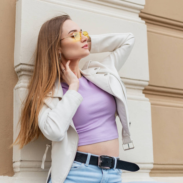 Beauty fashion street woman model with yellow vintage sunglasses in white leather jacket, purple r-shirt and jeans sits and poses near a vintage building on the street - Φωτογραφία, εικόνα