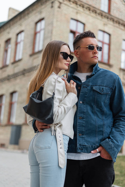 Fashion street beautiful young stylish couple guy and woman hipster with vintage sunglasses in fashionable denim clothes with a purse walking together in the city - Fotó, kép