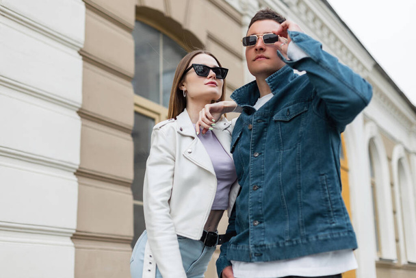 Cool fashionable young beautiful couple girl and boy with sunglasses in fashion casual outfit with denim jacket and leather jacket walks in the city. Trendy stylish clothes looks - Zdjęcie, obraz