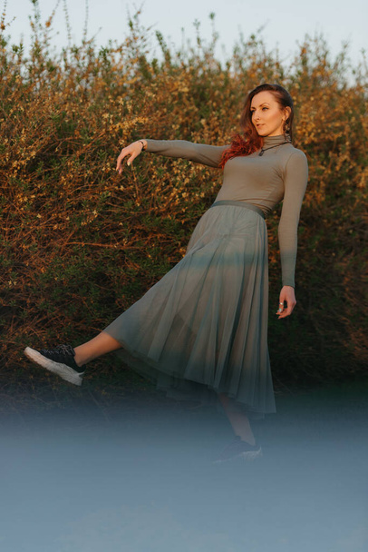 A happy redhead woman in green clothes is dancing in the park at sunset. - Photo, Image