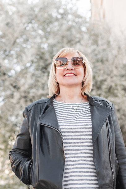 Fashion happy beauty elderly woman with a smile and trendy sunglasses in a stylish leather jacket and striped T-shirt walking in the city with a blooming tree - Photo, image